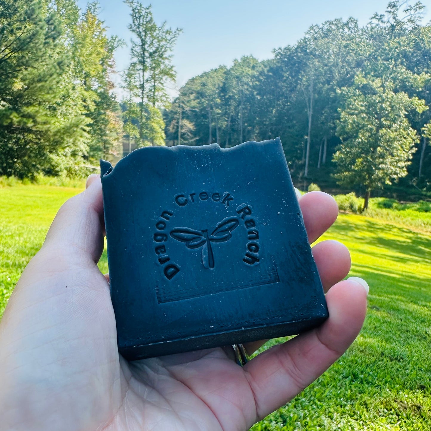 Hand holding a black soap bar with 'Dragon Creek Ranch' branding against a green outdoor background