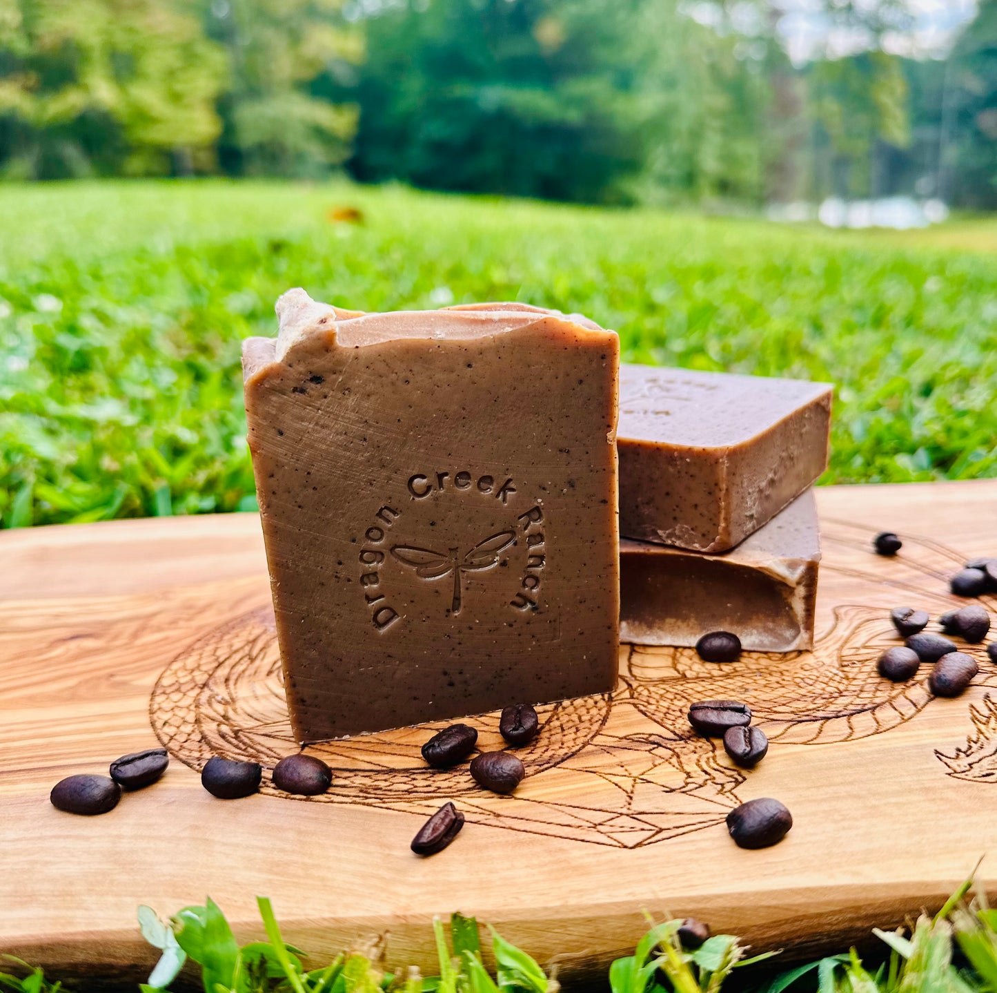 Three bars of soap with a dragon creek ranch logo on a wooden board outdoors. with scattered coffee beans on the board.