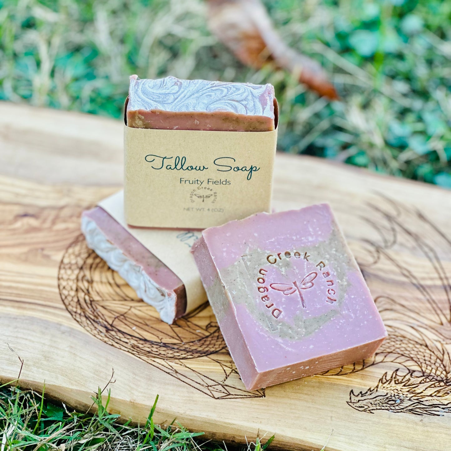 Three bars of pink and green colored soap labeled 'Tallow Soap' on a wooden surface with a natural background.