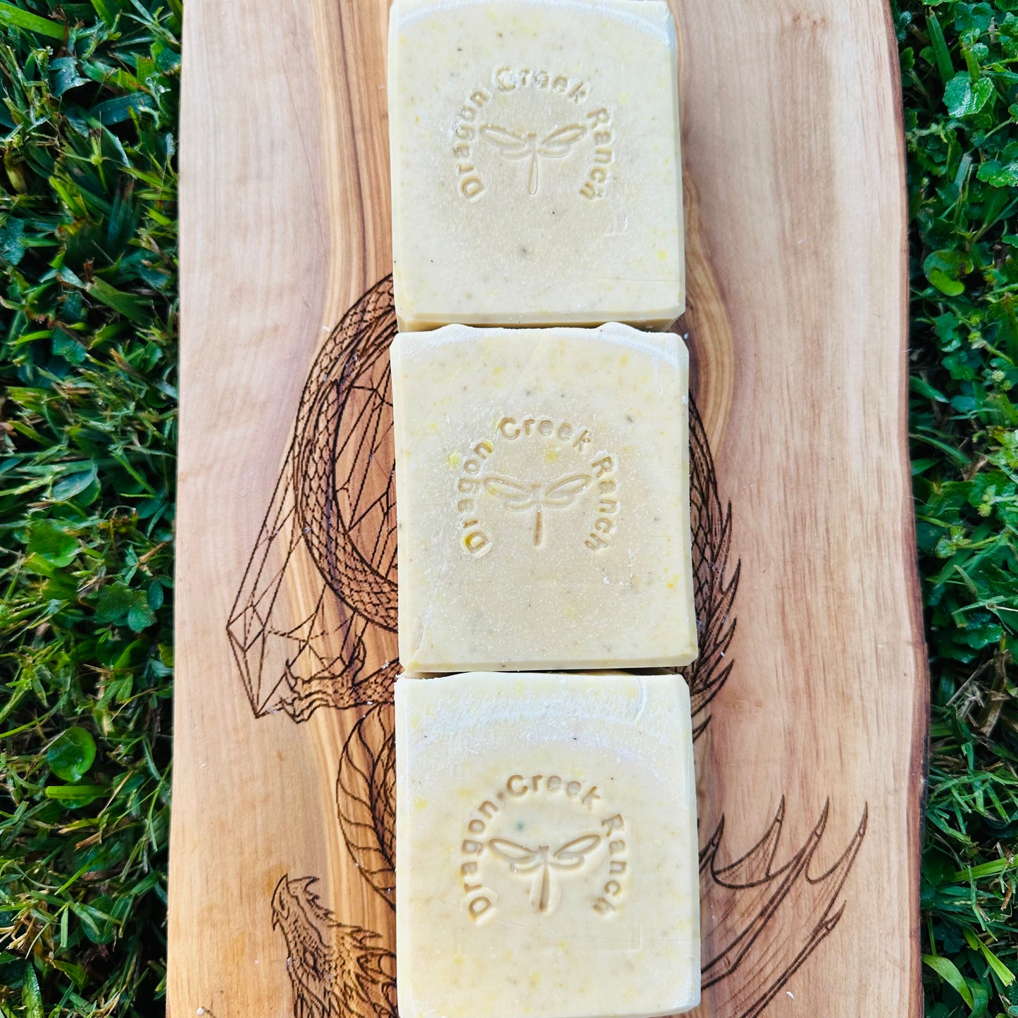 Three bars of soap with 'Dragon Creek' branding on a wooden board against a grassy background.