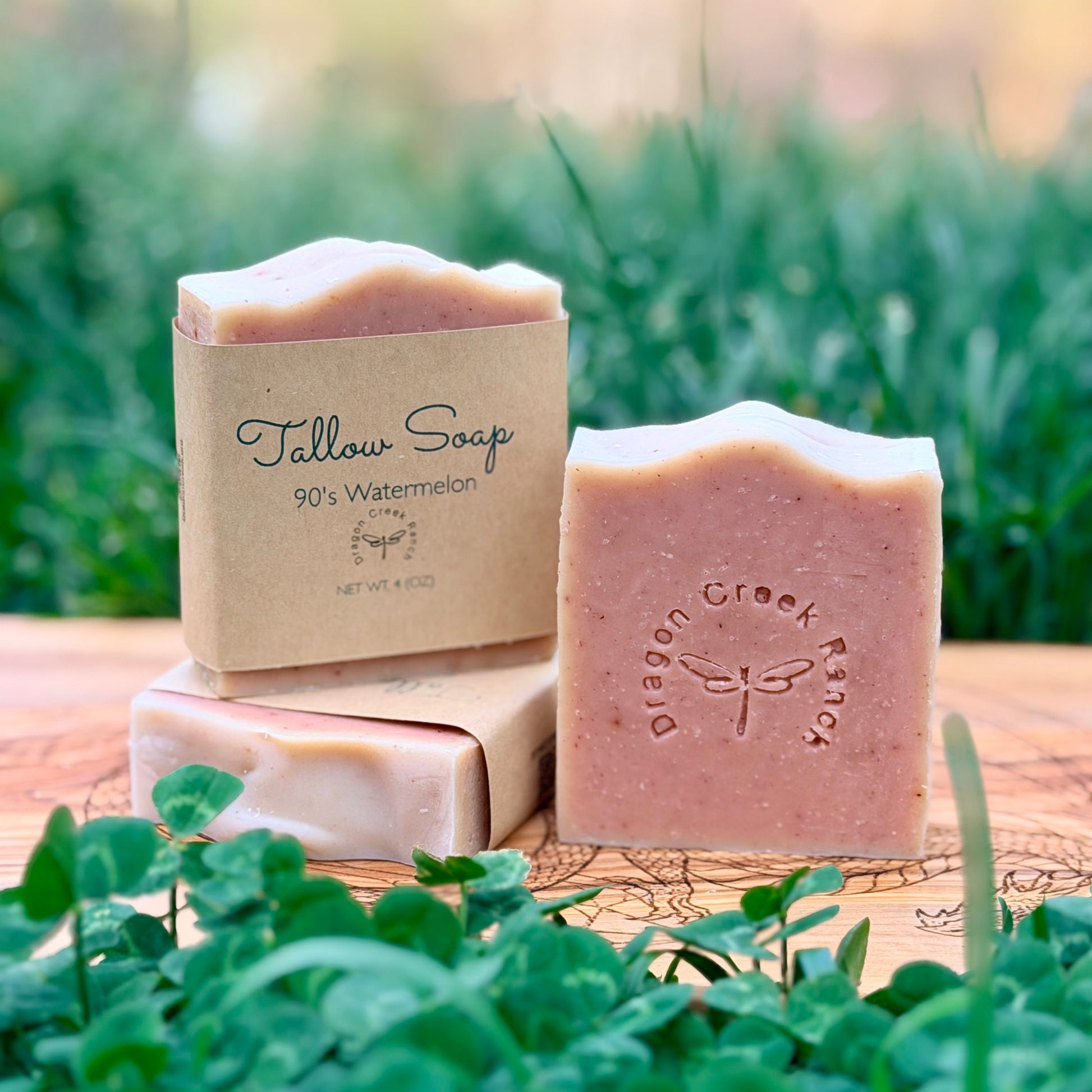 Three bars of pink soap and their packaging on a wooden surface with greenery in the background