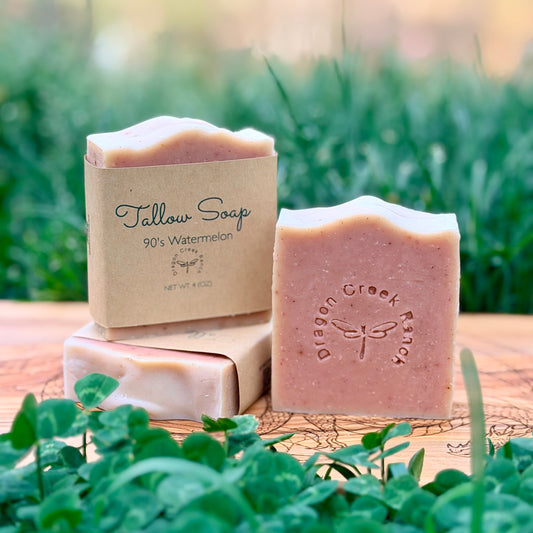 Three bars of pink soap and their packaging on a wooden surface with greenery in the background