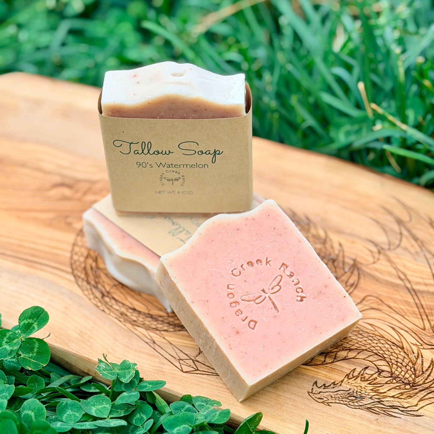 Three bars of pink soap labeled 'Tallow Soap' on a wooden surface with grass in the background.