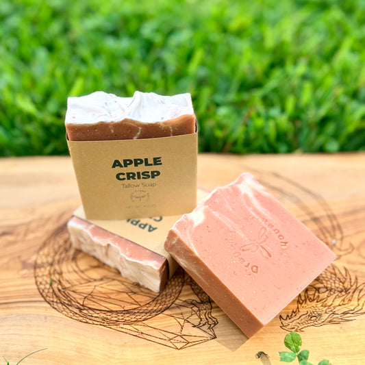 Three bars of handcrafted tallow soap displayed on a wooden board outdoors with a blurred green grass background. One bar is in a kraft paper sleeve labeled 'APPLE CRISP Tallow Soap', and two bars are loose, showing a dusty rose pink and cream marbled texture.