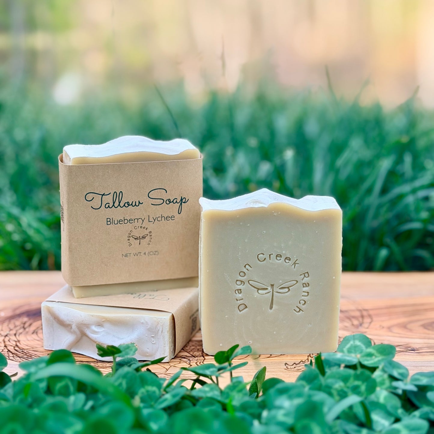 Three bars of soap, two wrapped in brown paper and the other unwrapped, with a Dragon Creek Ranch stamped on the soap, on a wooden surface with a natural background.