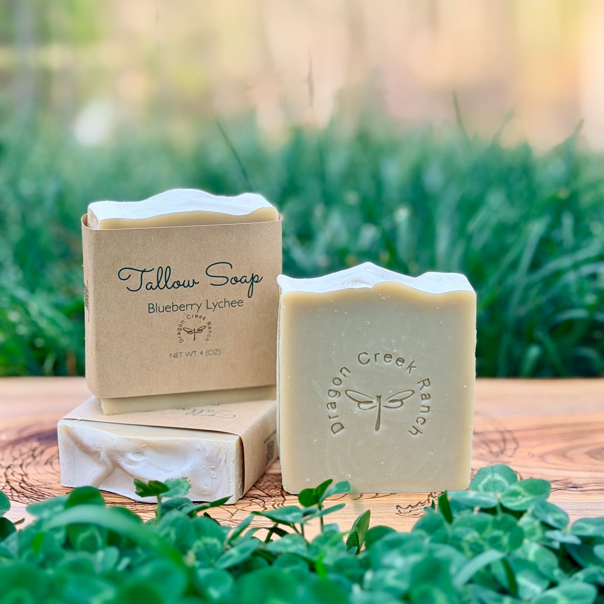 Three bars of soap, two wrapped in brown paper and the other unwrapped, with a Dragon Creek Ranch stamped on the soap, on a wooden surface with a natural background.