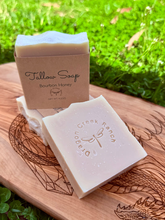 Three bars of soap labeled 'Tallow Soap' on a wooden board with grass in the background.