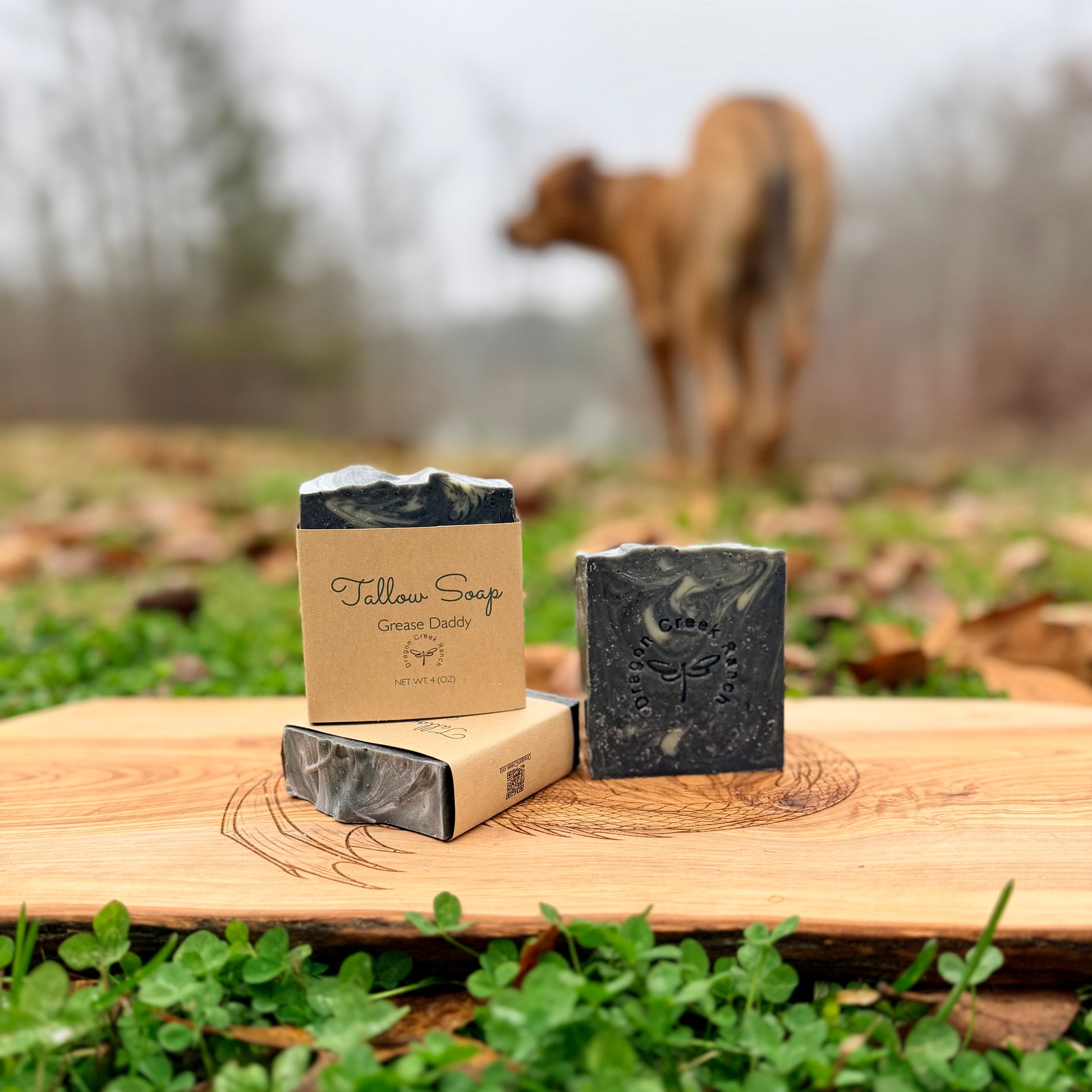 Three bars of soap and their packaging on a wooden surface with a blurred natural background containing a dog