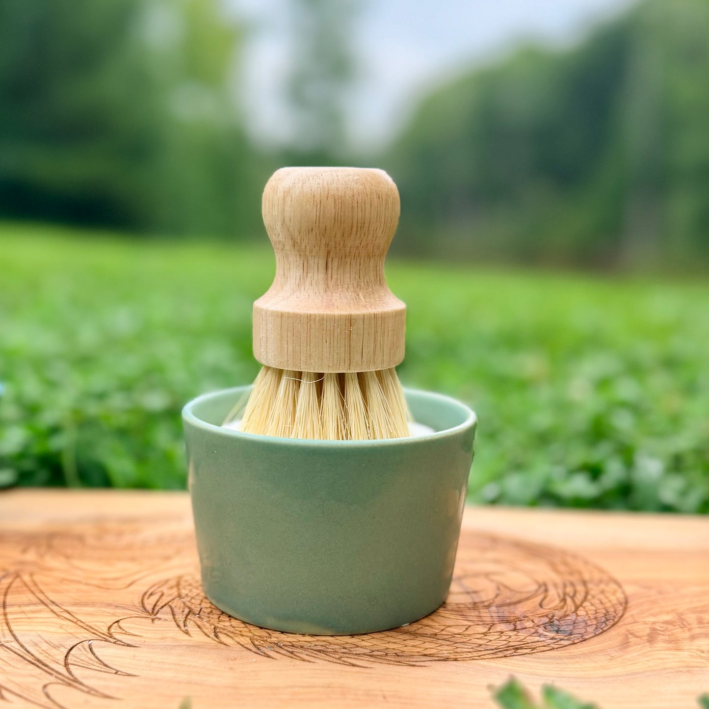 A small cleaning brush with a light wood handle and natural bristles sits in a pale green ceramic cup, placed on a wooden surface with a blurred green outdoor background.