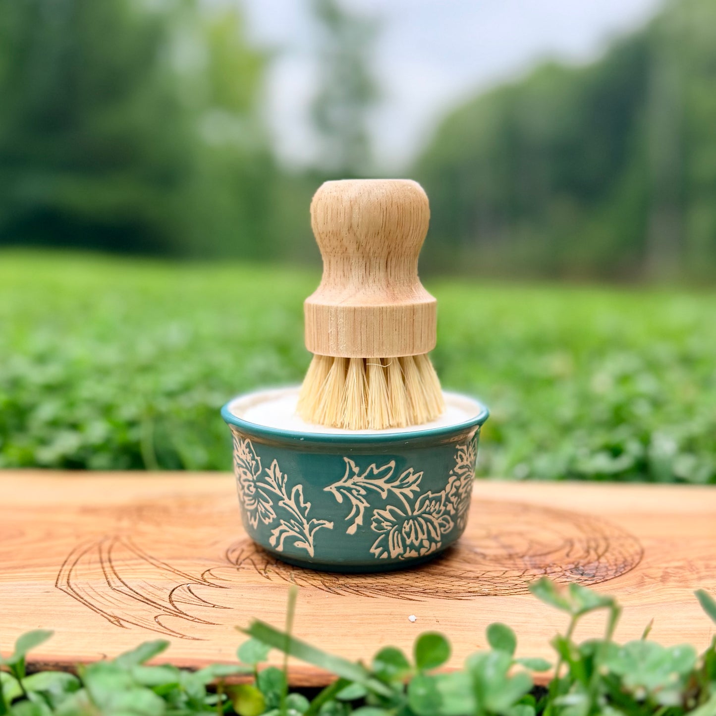 A small cleaning brush with a light wood handle and natural bristles sits in a teal ceramic cup with a white floral pattern, placed on a wooden surface outdoors with blurred green grass and trees in the background.