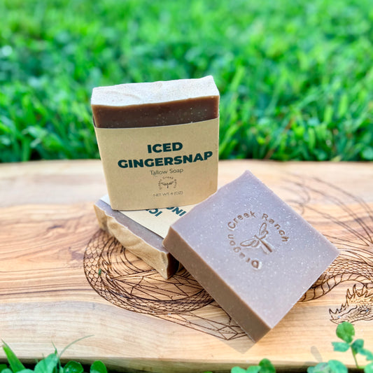 Three bars of soap on a wooden board with a blurred green grass background. One bar is wrapped in a kraft paper sleeve labeled 'ICED GINGERSNAP', and the other is loose, showing its brown color and embossed logo.
