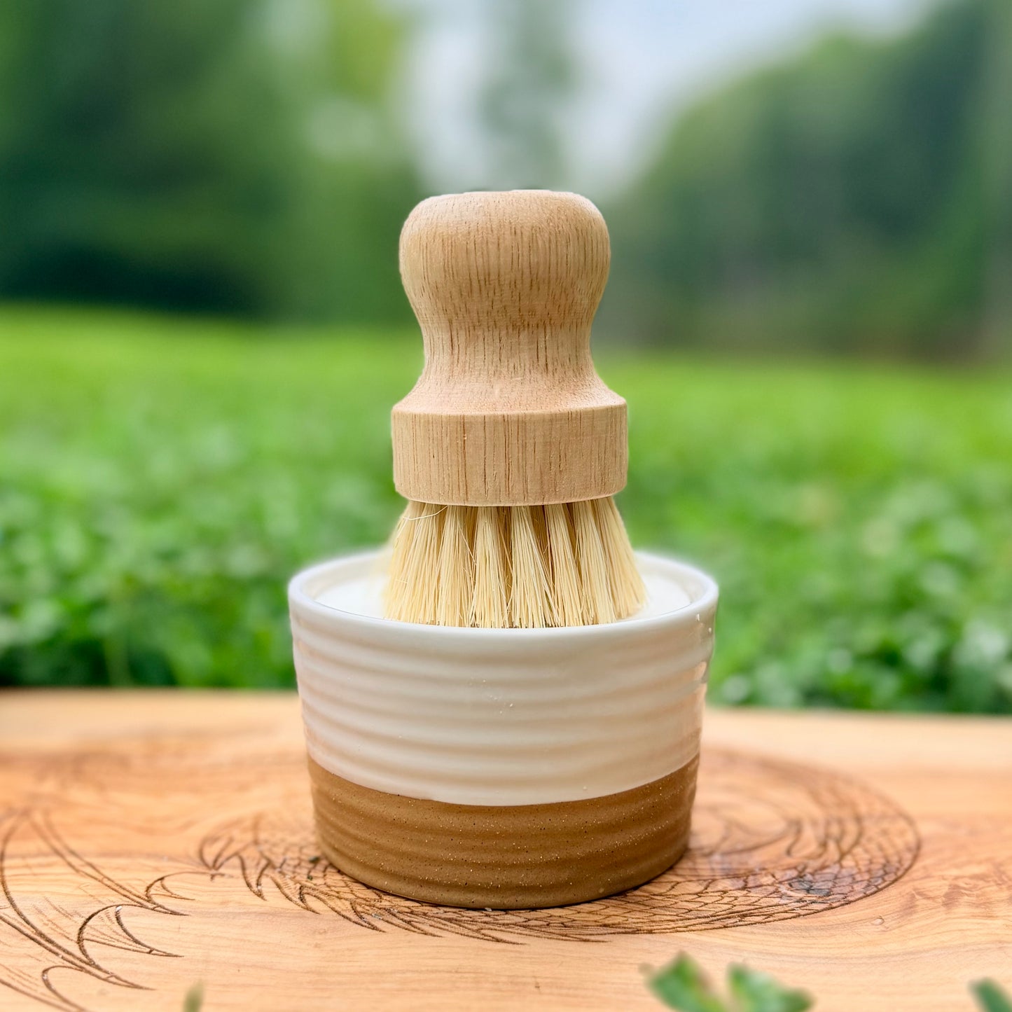 A wooden-handled cleaning brush with natural bristles and a two-tone ceramic base, set on a wooden surface with a blurred green outdoor background.