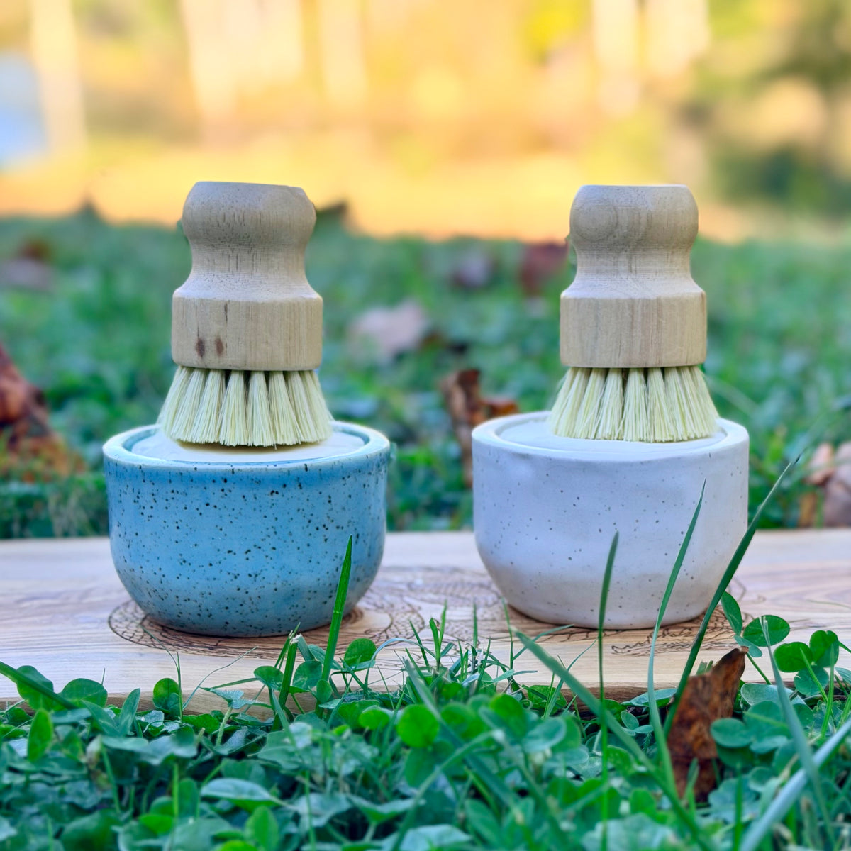 Blue and white speckled pottery ramekins side by side, each holding a wooden dish brush, on a board in the grass