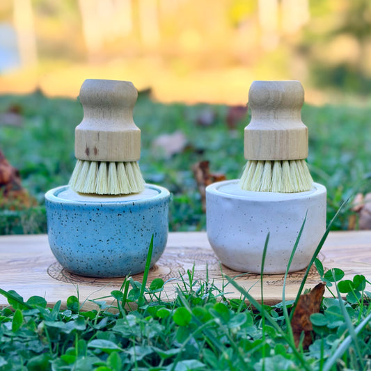 Blue and white speckled pottery ramekins side by side, each holding a wooden dish brush, on a board in the grass