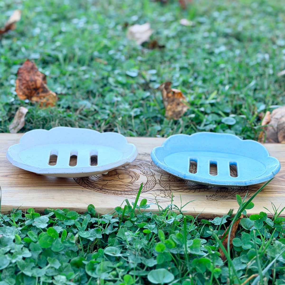 White and blue scalloped pottery soap trays with three drain slots, side view on a wood board in the grass