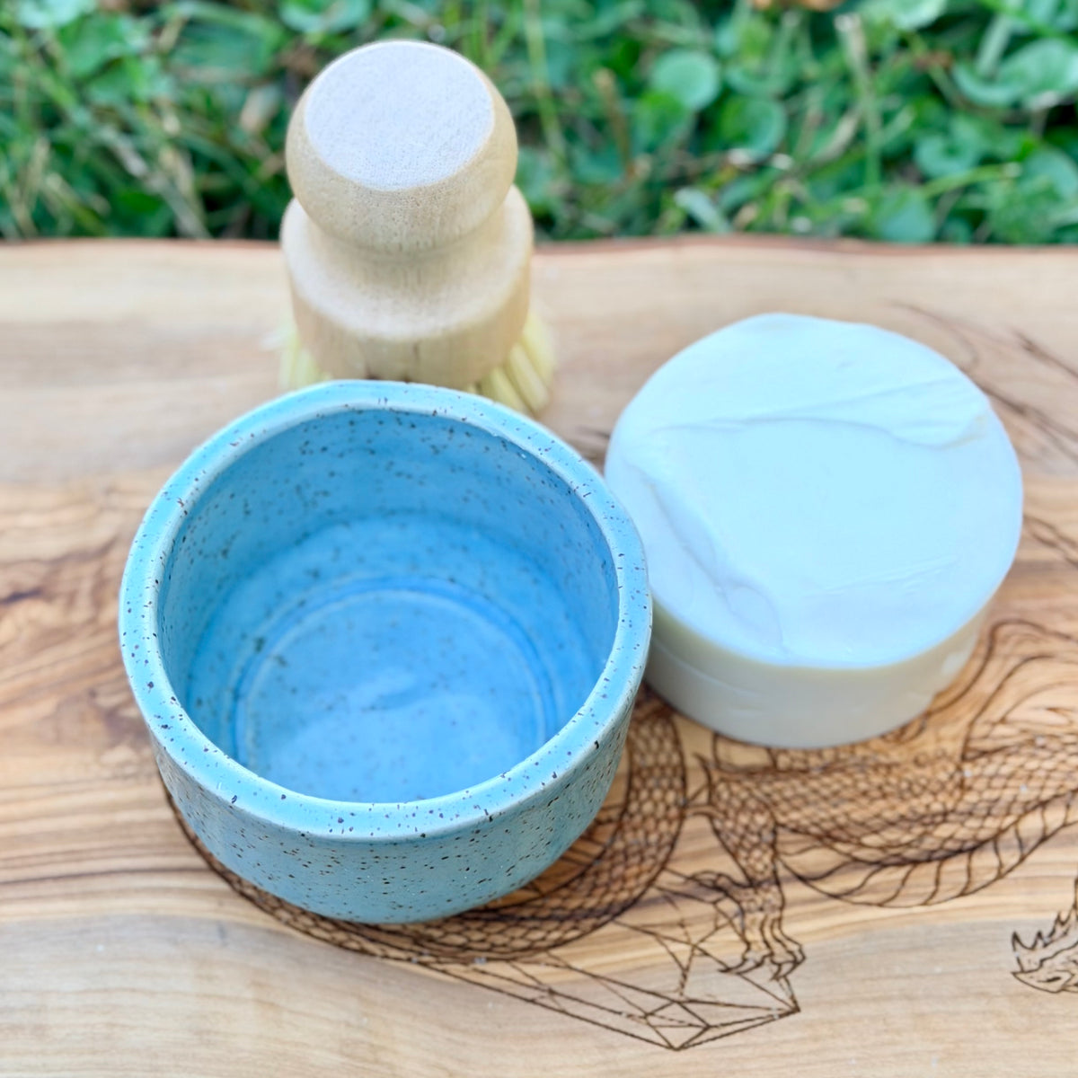 Open blue speckled pottery ramekin beside a round white tallow dish soap puck and a wooden dish brush