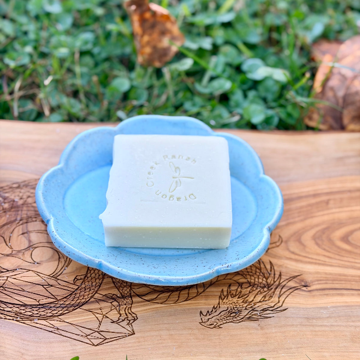 Blue scalloped pottery soap tray holding a cream stamped tallow soap bar on a dragon engraved wood board