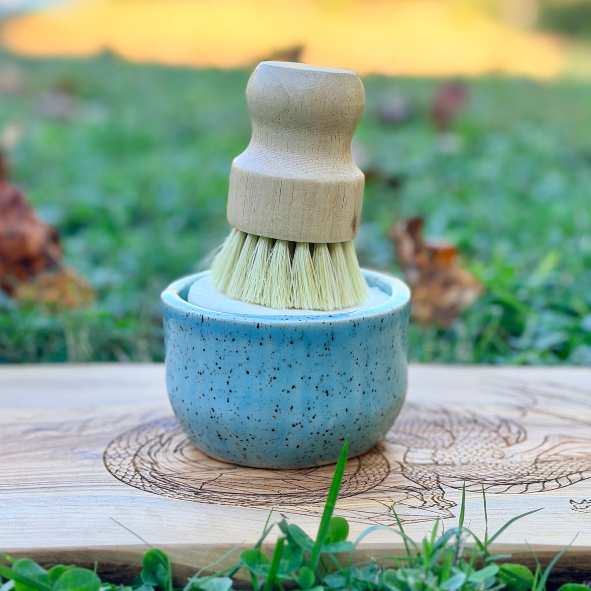 Blue speckled handmade pottery ramekin holding a wooden dish brush on a dragon engraved wood board outdoors