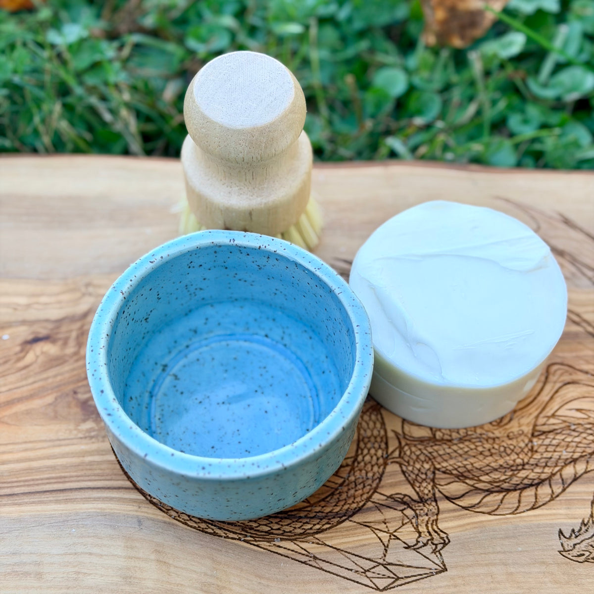 Empty blue speckled ramekin from above next to a white tallow dish soap puck and wooden brush on an engraved board