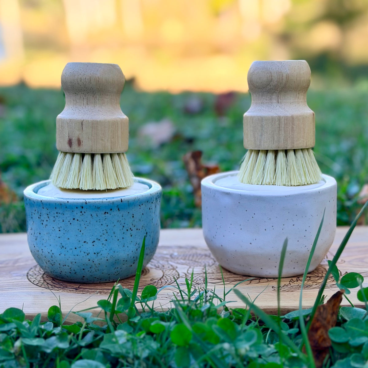 Blue and white handmade ramekin dish sets with wooden brushes on a wood board in green grass with fall leaves