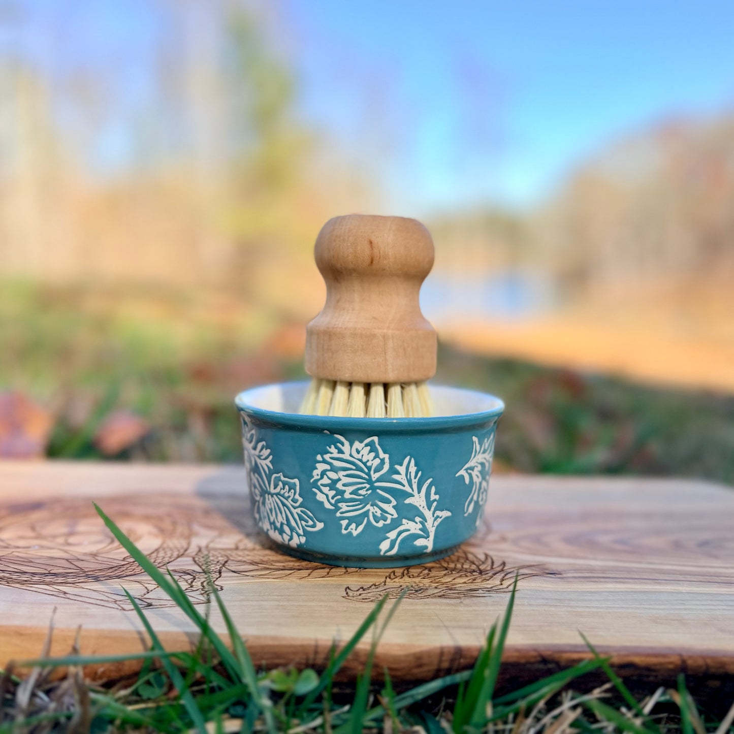 Cleaning brush with wooden handle and floral-patterned ramekin on a wooden surface outdoors.