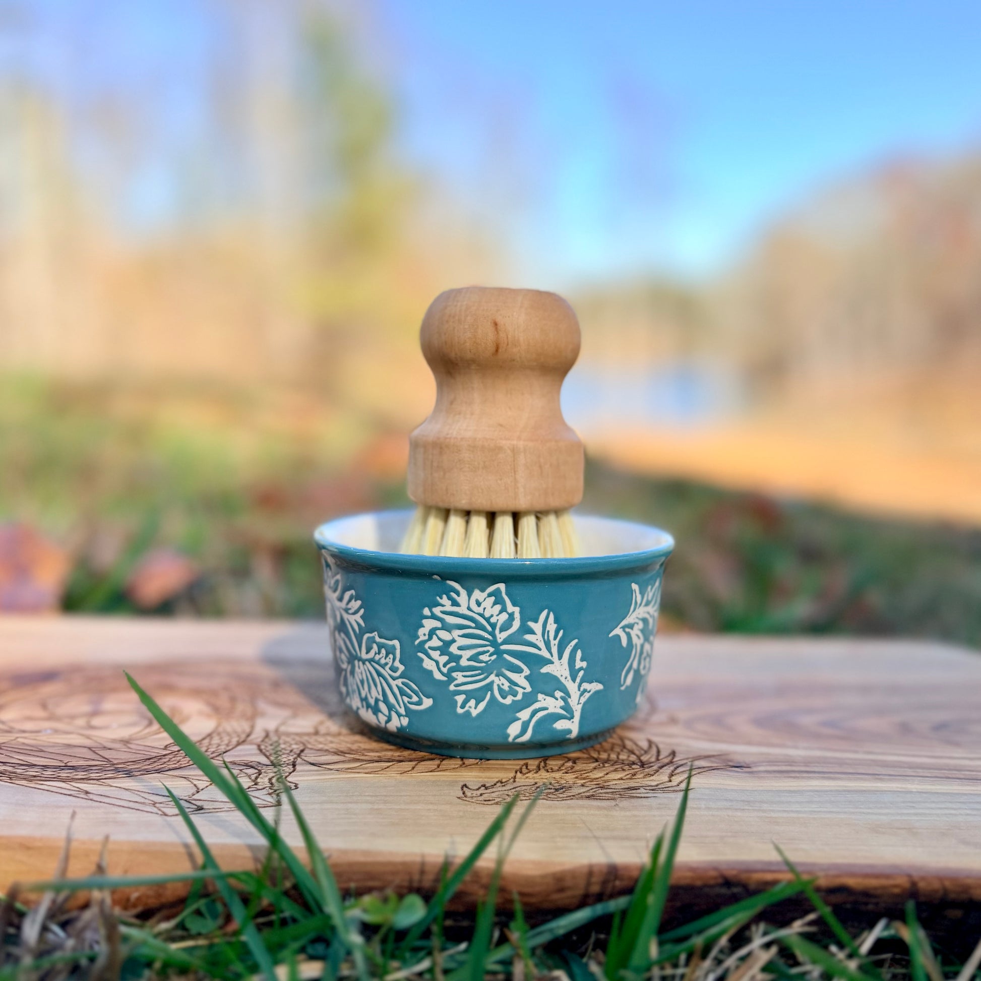 Cleaning brush with wooden handle and floral-patterned ramekin on a wooden surface outdoors.