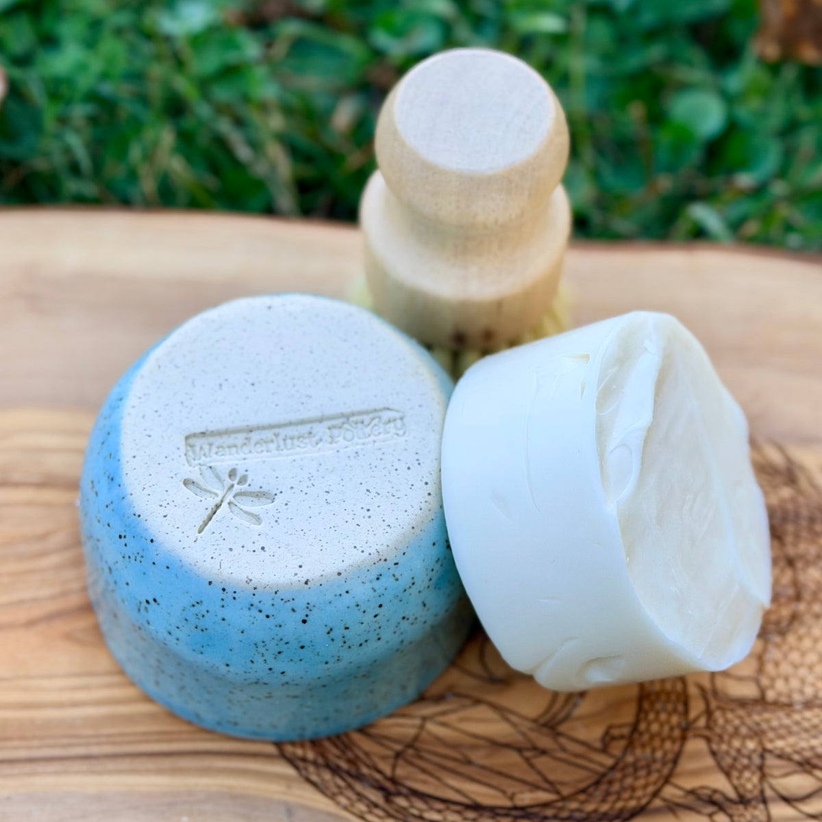 Wanderlust Pottery dragonfly stamp on an upturned blue ramekin with a white dish soap puck and wooden brush