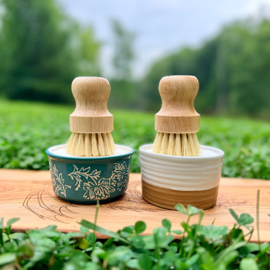 Two small hand brushes with light wood handles and natural bristles sit in small ceramic bowls on a wooden surface outdoors, with green plants and a blurred garden background.
