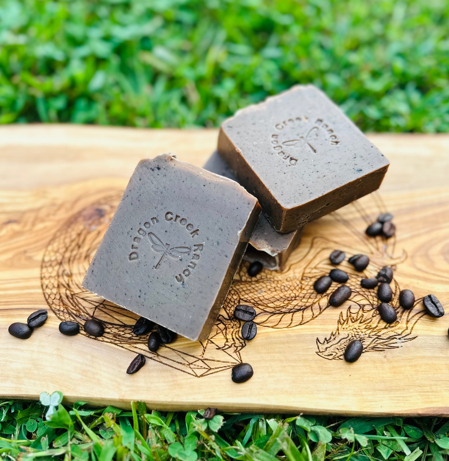 Three pieces of dark brown soap on a wooden board with scattered coffee beans, outdoors.