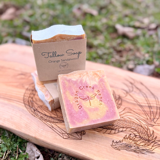 Three bars of soap, one in a package labeled 'Tallow Soap' and the other with 'Dragon Creek Ranch' branding, on a wooden board outdoors.
