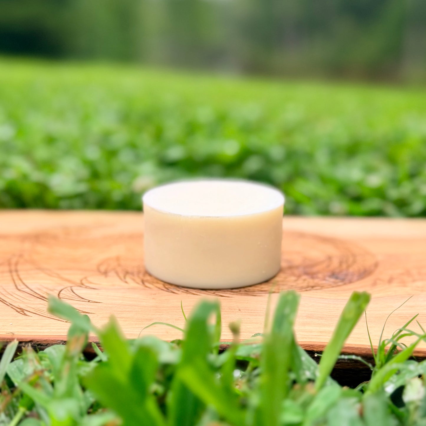 A small round white/beige soap sits on a wooden board outdoors, with blurred green grass in the background and foreground.
