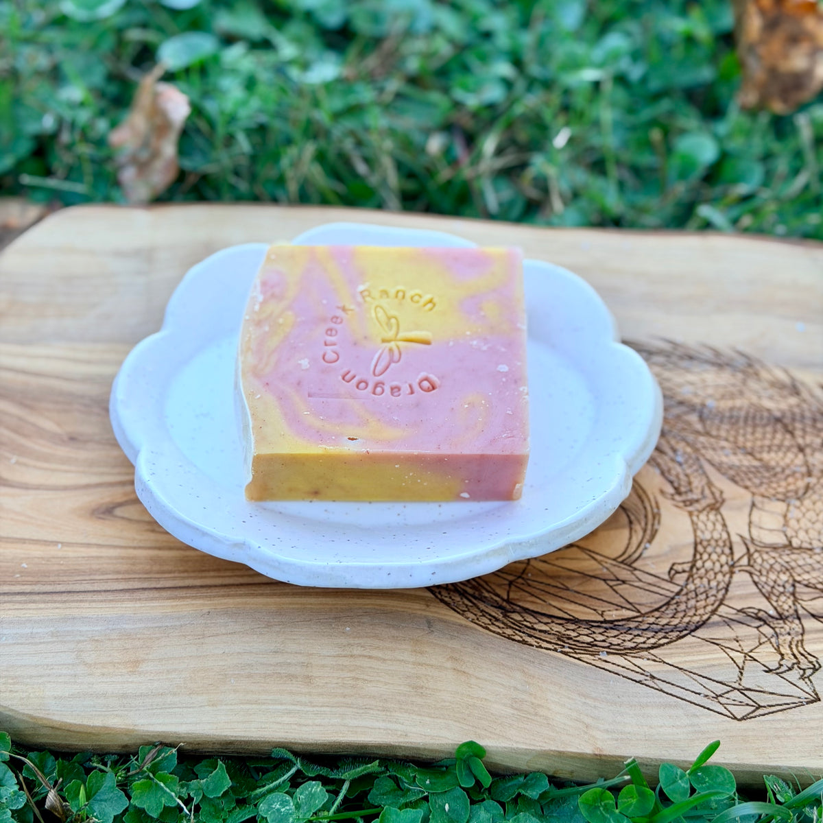 White scalloped pottery soap tray holding a pink and yellow swirled stamped tallow soap bar on an engraved board