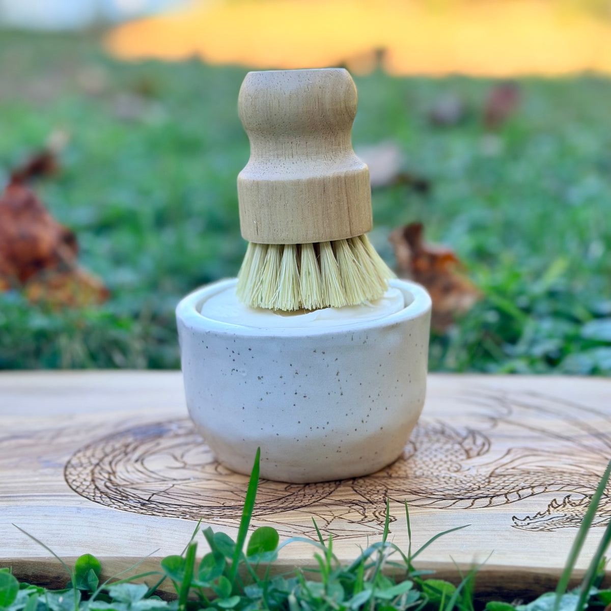 White speckled handmade pottery ramekin holding a wooden dish brush on a dragon engraved wood board outdoors